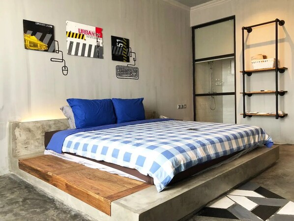 A bedroom with a bed, blue pillows, a checkered bedspread, a wooden platform, a wall-mounted shelf, and a mirror.