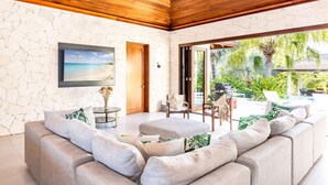 A modern living room with a wooden ceiling, a large window with a view of a tropical garden, and a comfortable seating area. - Villa Khaya - Bespoke Waterfront Home (Providenciales)