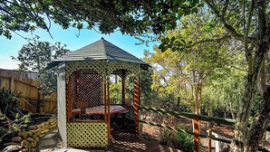 Un gazebo in legno con tetto a graticcio e una panca all'interno, circondato da alberi e un sentiero in pietra.