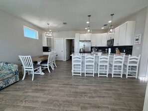 A modern kitchen with white cabinetry, a central island with bar stools, and a dining area with a wooden table and white chairs. - Shared Heated Pool, Pickleball Court, Pet Friendly, Golf Cart Access (Port Aransas)
