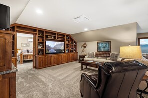 A spacious living room with a large flat-screen TV, wooden shelving, a sofa, and a leather chair.