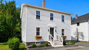 A two-story white house with a front porch, a small front yard, and a clear blue sky. - The Coven House (Salem)