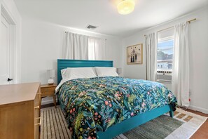 Un dormitorio con cama color turquesa, cortinas blancas, un cuadro en la pared y vistas al exterior a través de una ventana.
