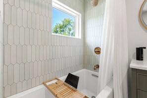 Un baño con bañera, ventana con vista a árboles y zona de ducha.