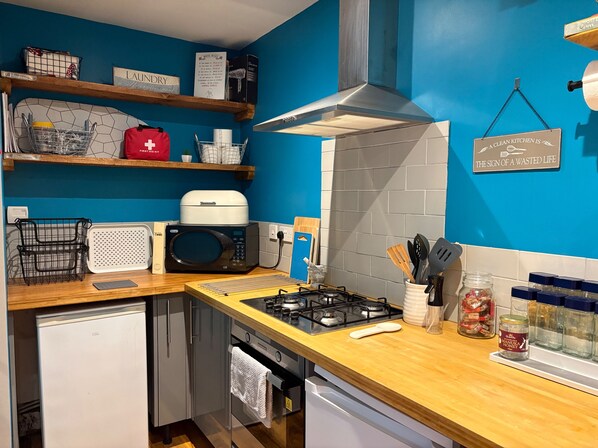 A compact kitchen with a microwave, stove, and a small refrigerator. There are shelves with kitchen items and a sign on the wall.