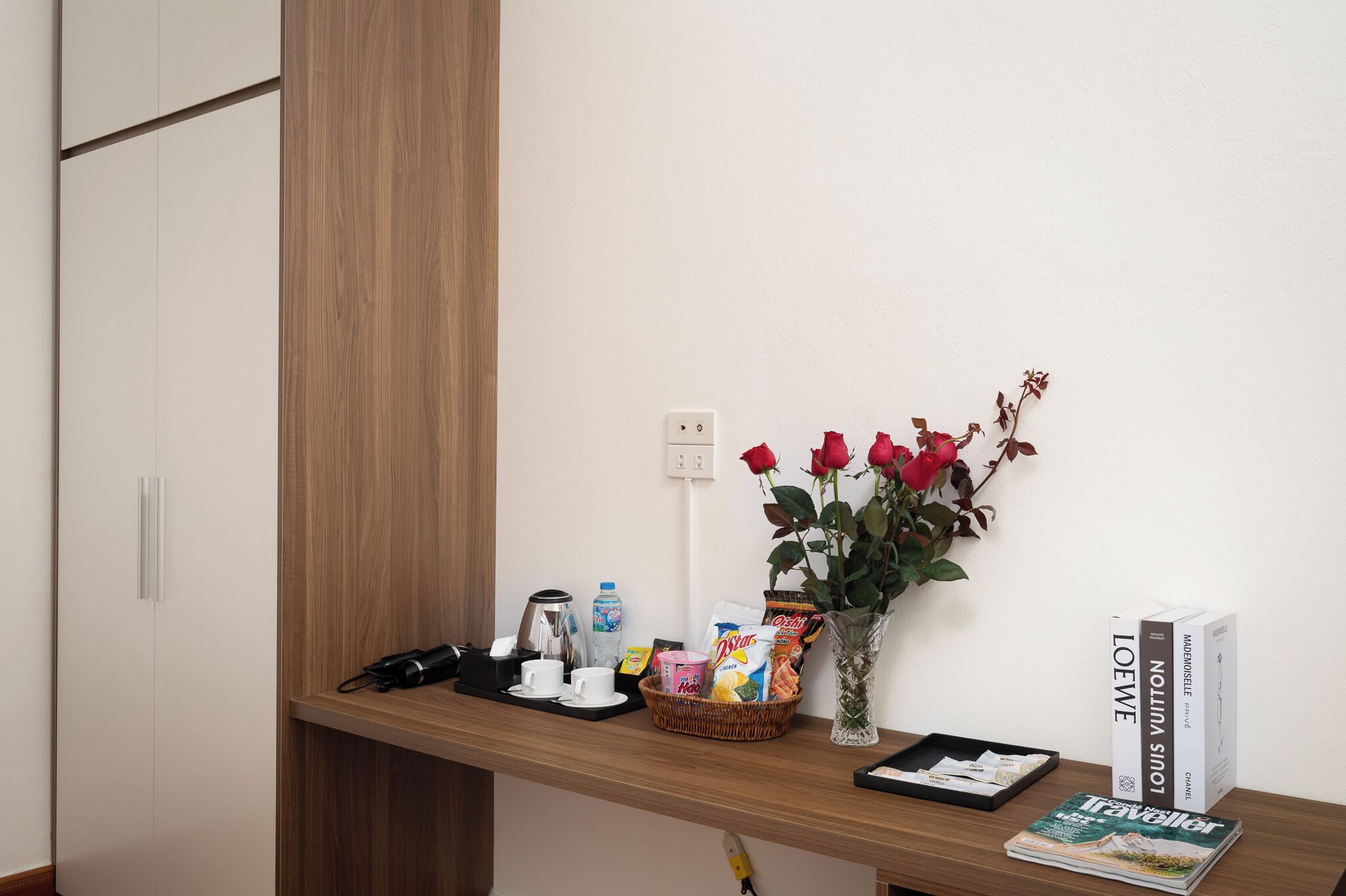 A wooden desk with a basket of snacks, a coffee maker, a vase with red roses, and a stack of books.