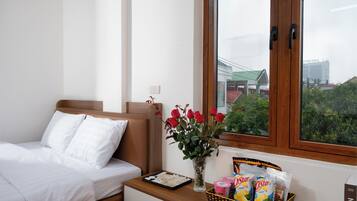 A hotel room with a bed, a nightstand with a vase of red roses, a basket of snacks, and a window with a view of buildings and greenery.