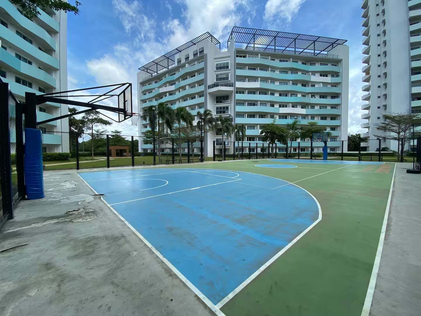 An outdoor basketball court with a blue and green surface, surrounded by a fence and palm trees, with high-rise buildings in the background.