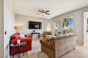 A living room with a red couch, a brown couch, a ceiling fan, a TV, and a view of palm trees through large windows.