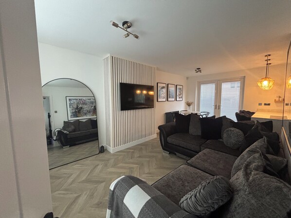 A modern living room with a dark grey sofa, a flat-screen TV mounted on the wall, and a wooden herringbone floor. - Charming house with sauna in Glasgow perfect for your getaway (Glasgow)