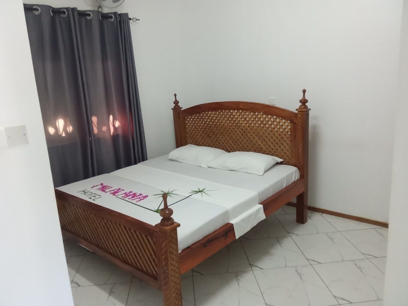 A single bed with a wooden frame and a white mattress, a dark curtain, and a white wall.