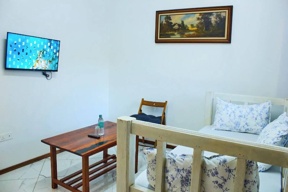 A room with a wooden table, a chair, a bed with floral bedding, a painting on the wall, and a TV displaying a map.