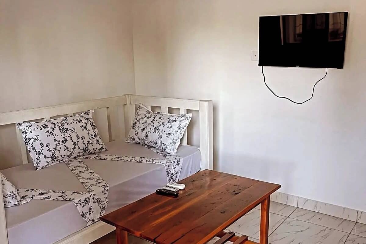 A room with a white bed, a wooden table, and a TV mounted on the wall.