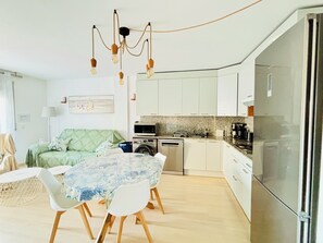 A compact kitchen with white cabinets, a stainless steel refrigerator, and a dining area with a table and chairs. - PORT CANIGO Duplex with private solarium (ROSES)