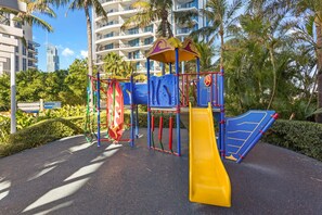 A playground with a yellow slide and climbing structures, surrounded by palm trees and modern buildings. - Chevron Renaissance Ocean & River Views (Surfers Paradise)