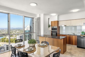 A modern kitchen with a view of a cityscape, a dining area with a table and chairs, and a large window. - Chevron Renaissance Ocean & River Views (Surfers Paradise)