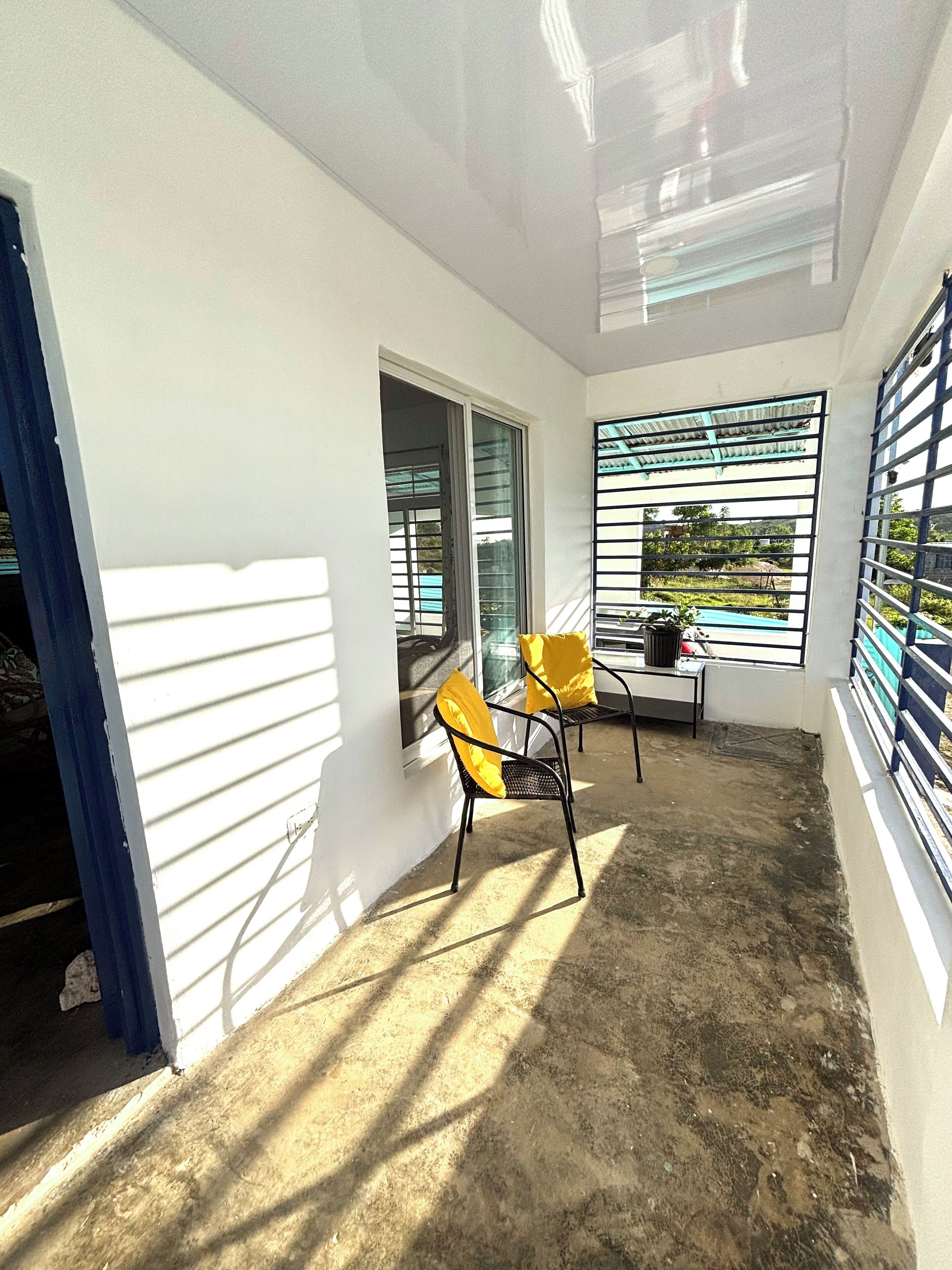 Un patio ensoleillé avec un sol en béton, deux chaises dotées de coussins jaunes et une table sur laquelle se trouve un objet noir.