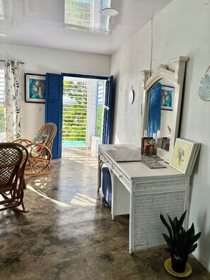 Une pièce avec un bureau blanc, des chaises en osier, une plante en pot et une vue sur l’extérieur, visible à travers une porte bleue.