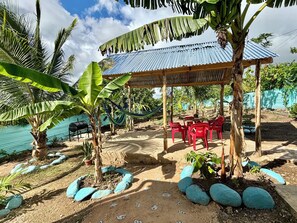 A covered outdoor seating area with red plastic chairs and tables, surrounded by tropical plants and rocks. - Terrific 2-bedroom house with AC in sunny Higüey (Higüey)