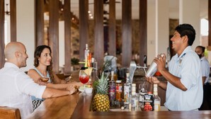 A bar area with a bartender preparing a drink, a couple seated at a table, and various bottles and glasses on the counter. - 3-bedroom Condo with AC, WiFi, Ocean Views, Tennis court & Gym in Vivo resorts (Bajos de Chila)