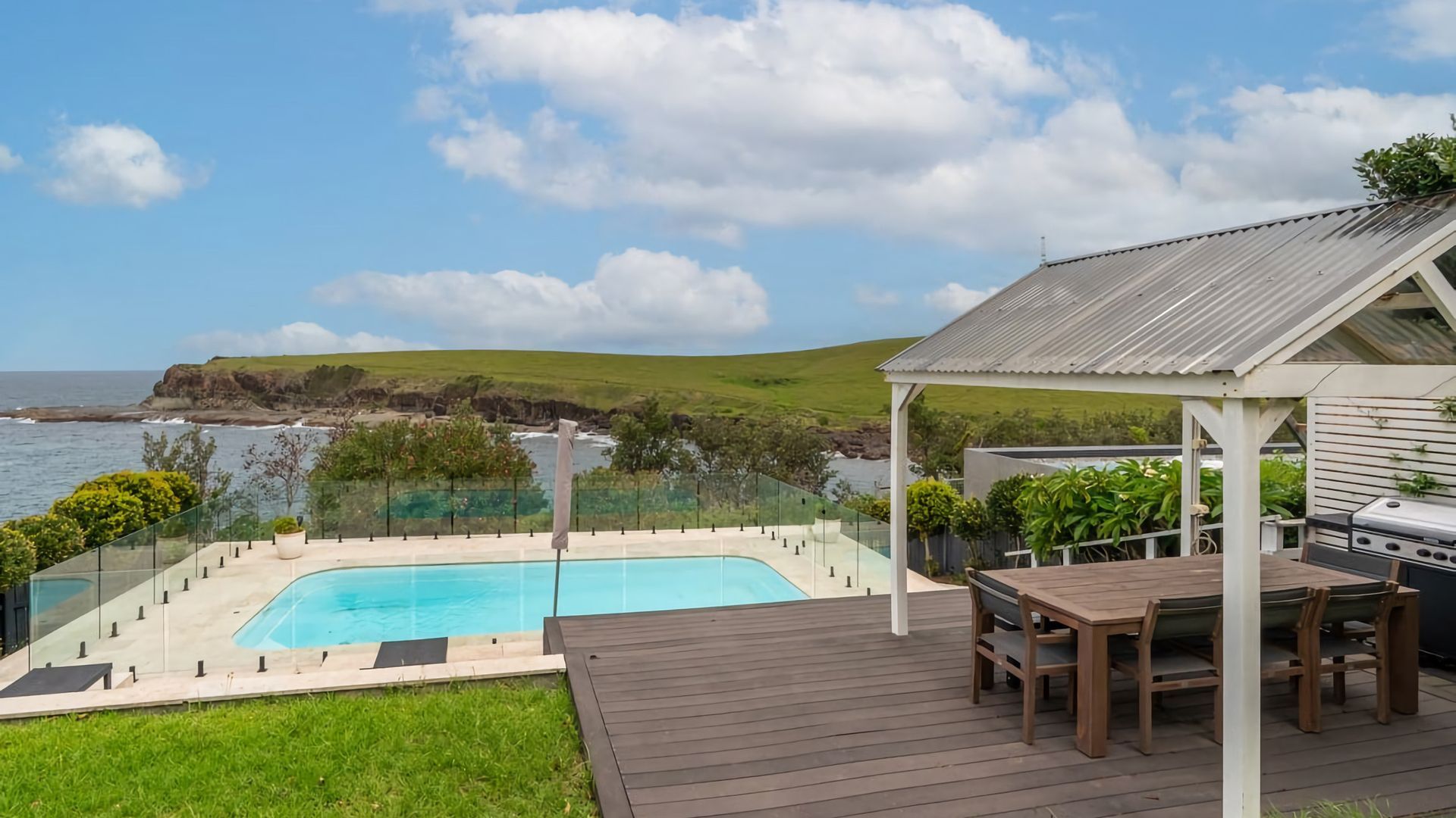 A pool with a view of the ocean, a wooden deck, and a barbecue area.