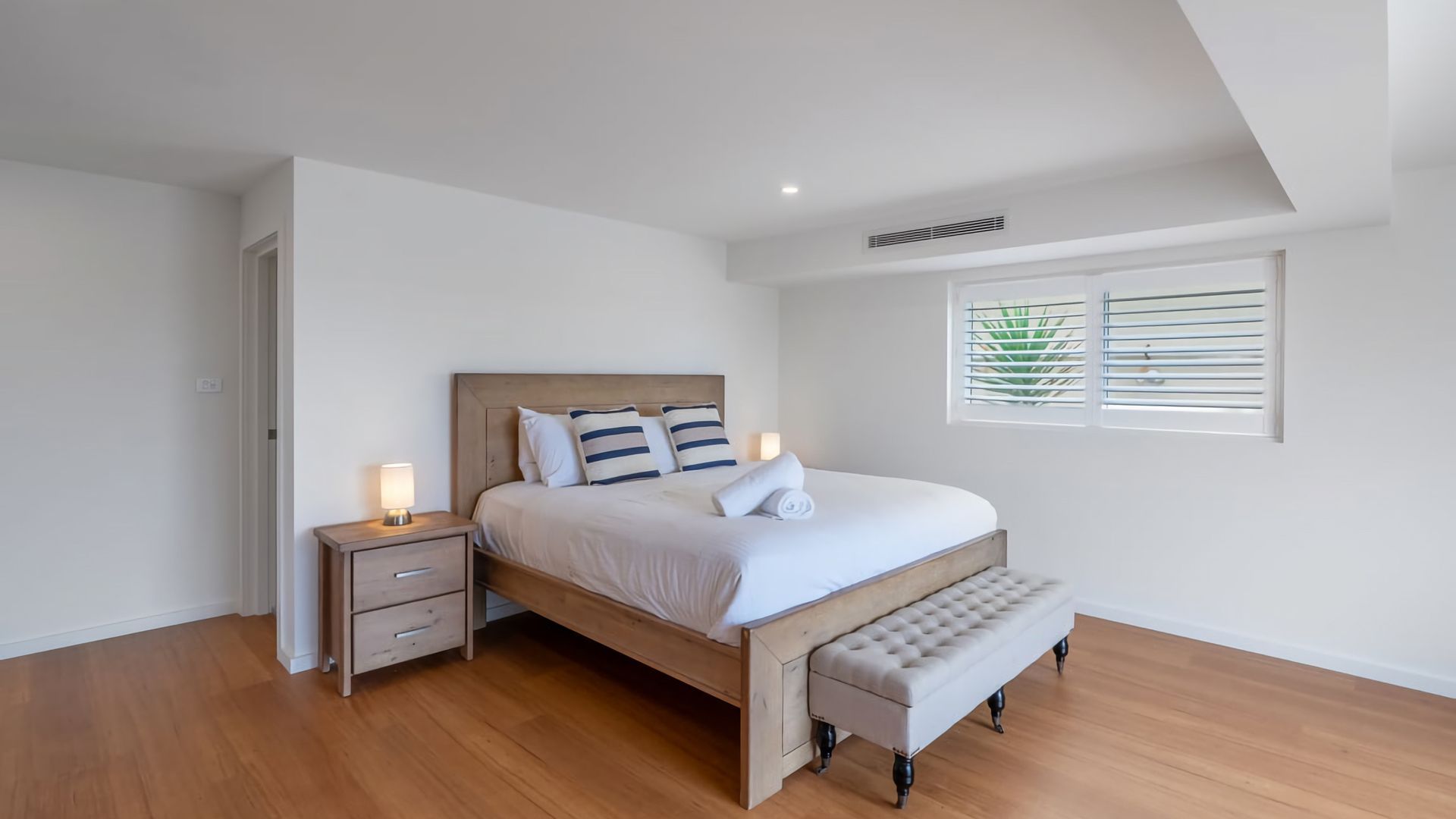 Une chambre à coucher avec un lit en bois, des tables de chevet, un banc et une fenêtre avec des stores.