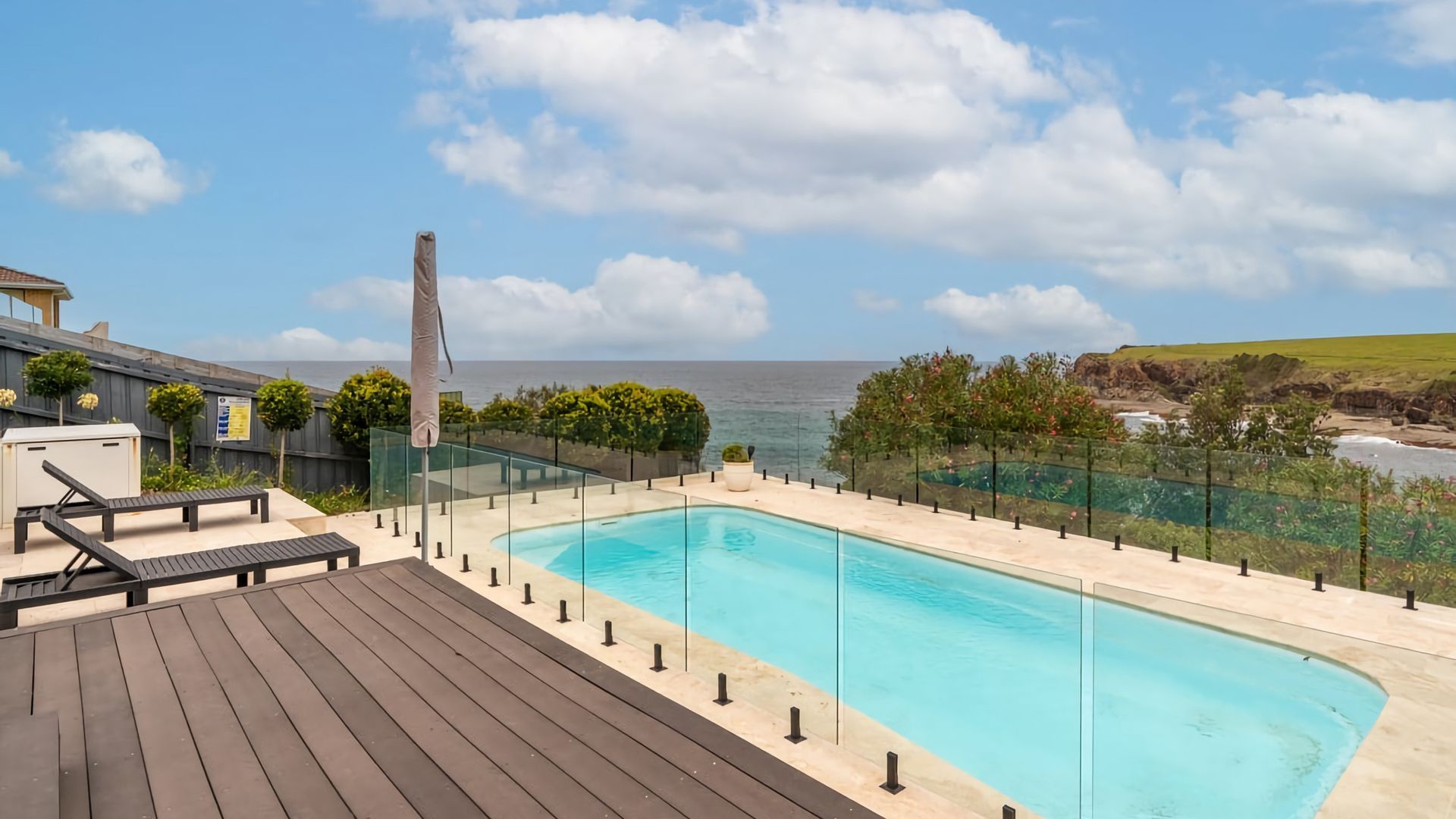 A swimming pool with a clear view of the ocean, surrounded by a wooden deck and glass railing.