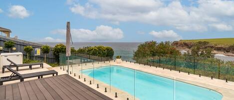 Une piscine offrant une vue dégagée sur l’océan, entourée d’une terrasse en bois et d’une balustrade en verre.