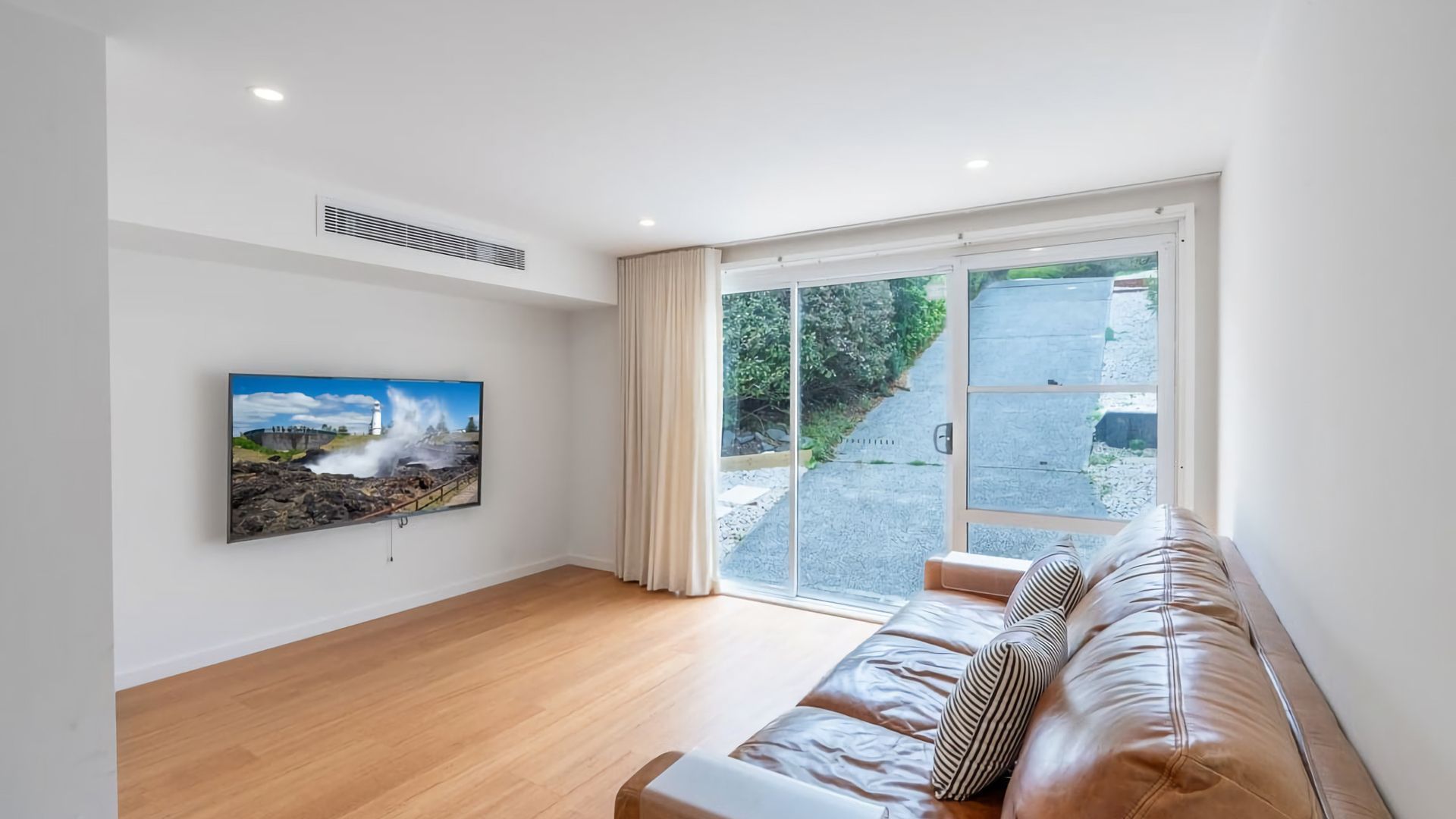A modern living room with a large window, a flat-screen TV mounted on the wall, and a leather sofa.