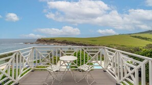 A wooden deck with white railings, a small table, and two chairs overlooking a coastal landscape with a grassy hill and ocean. - The Glenlorne: Your Serene Retreat Awaits (Kiama)