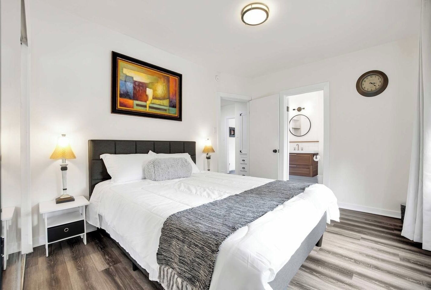 Ein ordentlich eingerichtetes Schlafzimmer mit einem Bett, Nachttischlampen, einem Gemälde, einer Uhr und einem durchgegangenen Tür erkennbaren Badezimmer.