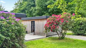 A wooden cabin with a door and steps leading to a porch, surrounded by lush greenery and flowering plants. - Beautiful home in Vejby with sauna (Vejby)