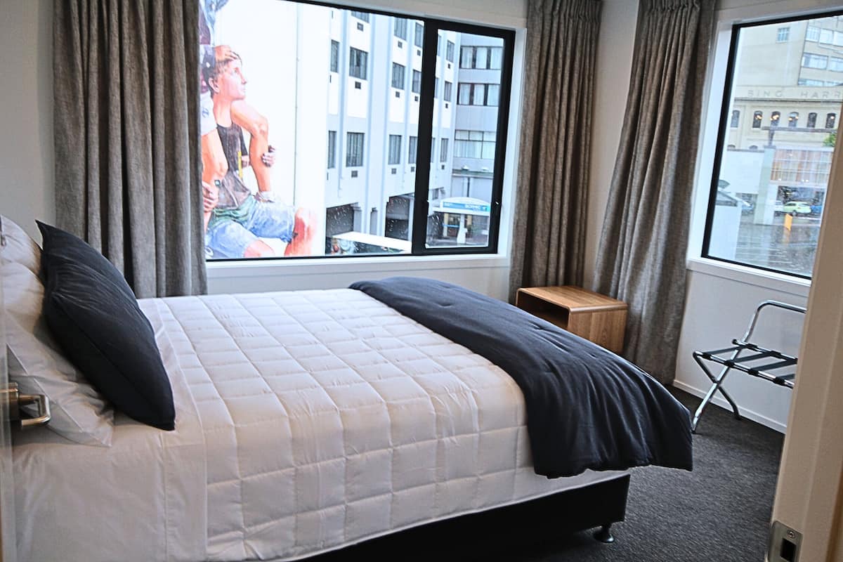 Una habitación de hotel con una cama, una ventana con cortinas, una mesita de noche de madera y una silla.