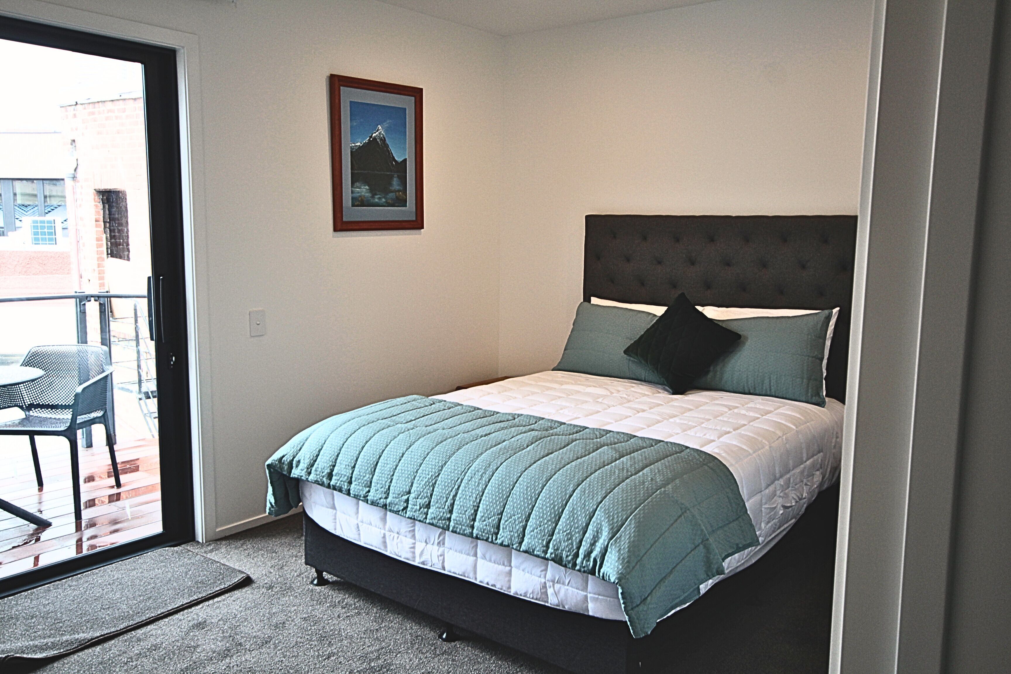 A bedroom with a bed, a framed picture on the wall, and a balcony with a chair.