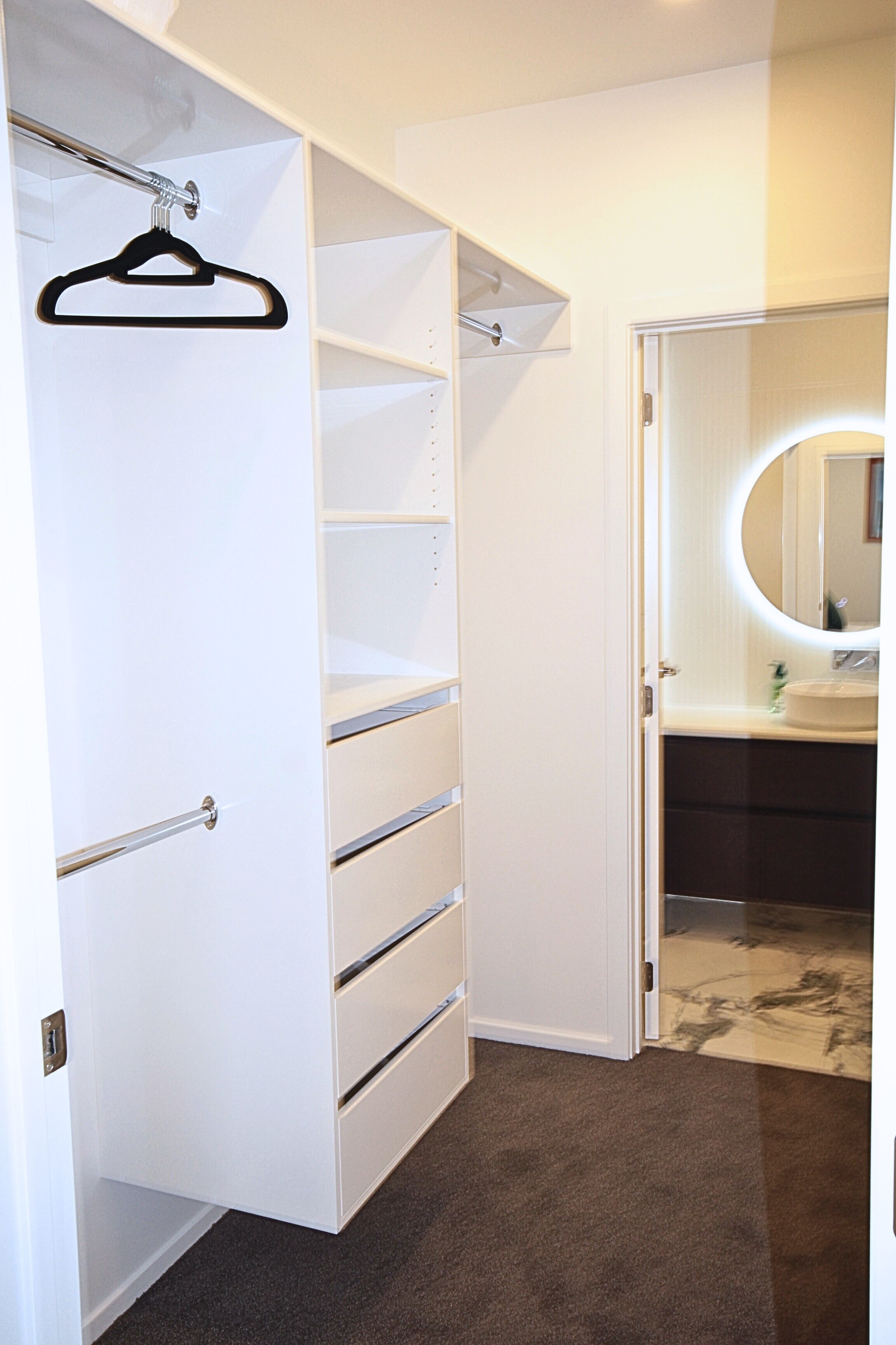 Un dressing sans porte, avec un portant à vêtements et une salle de bain dotée d’un miroir rond et d’un lavabo.