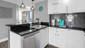 Een moderne keuken met witte kasten, zwarte aanrechtbladen en roestvrijstalen apparatuur. Er hangen hanglampen boven de gootsteen en een klok aan de muur.
