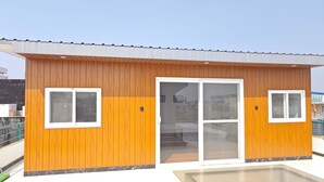 Ein kleines Holzgebäude mit zwei Fenstern und einer Schiebetür aus Glas, vermutlich ein Poolhaus oder ein Gerätehaus.