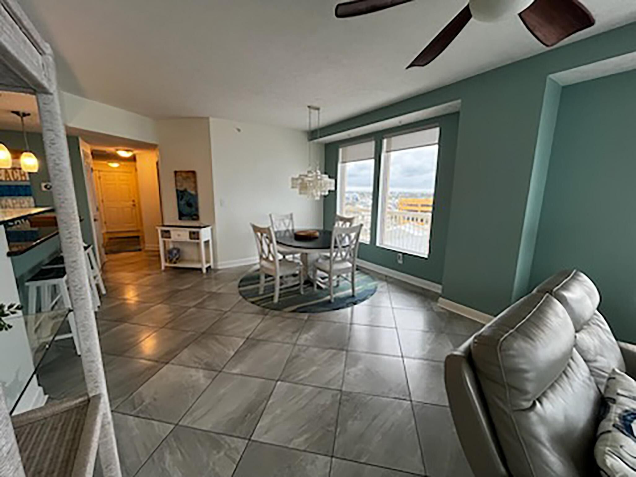 A spacious living room with a large window, a dining area with a round table, and a ceiling fan.