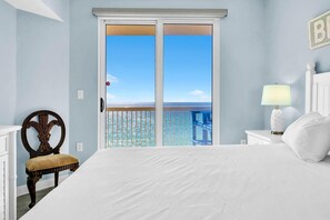 A bedroom with a large bed, a chair, a nightstand with a lamp, and a view of the ocean through a sliding door. - Seychelles 701 (Panama City Beach)
