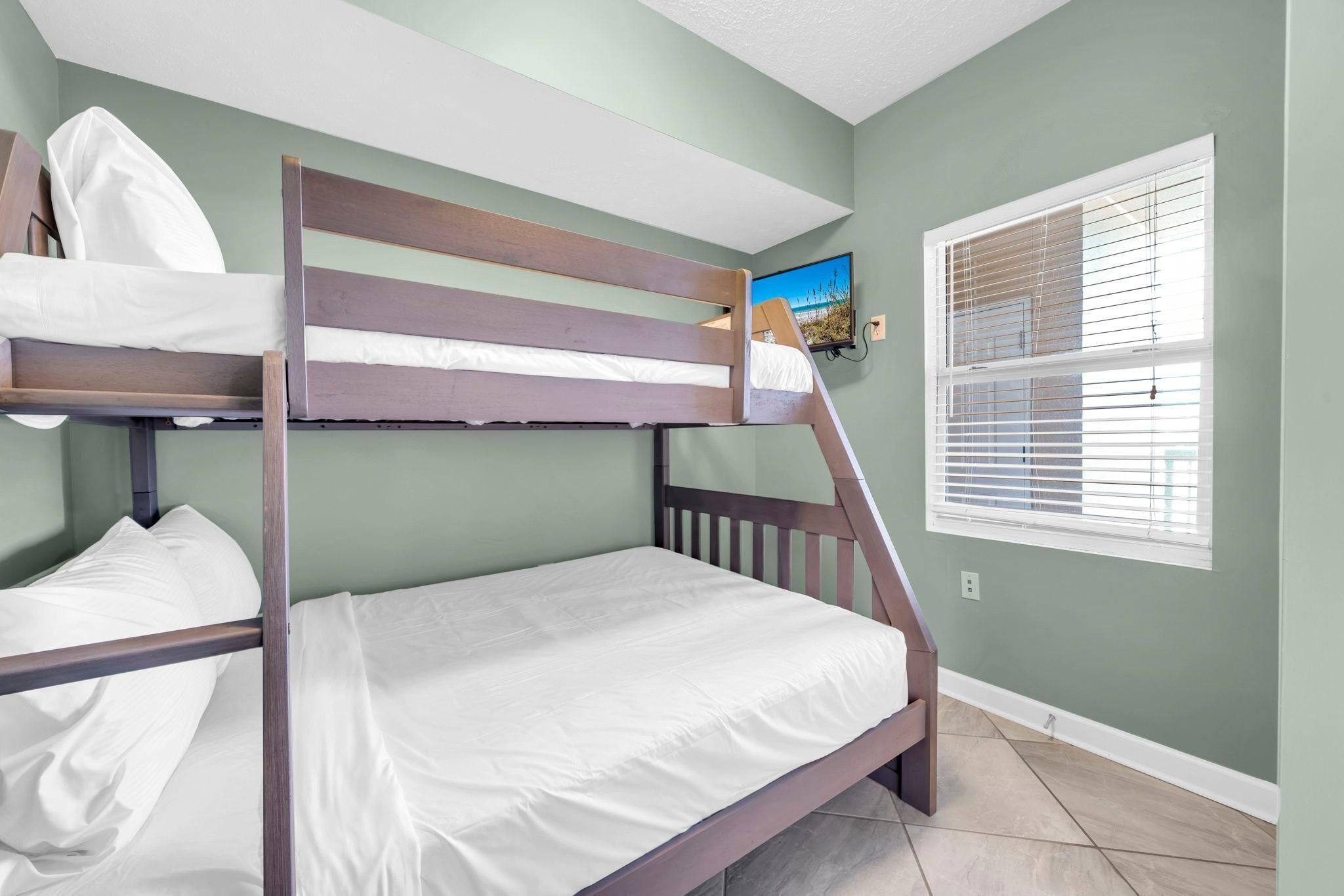 Una cama litera con ropa de cama blanca en una habitación con paredes verdes y una ventana con persianas.
