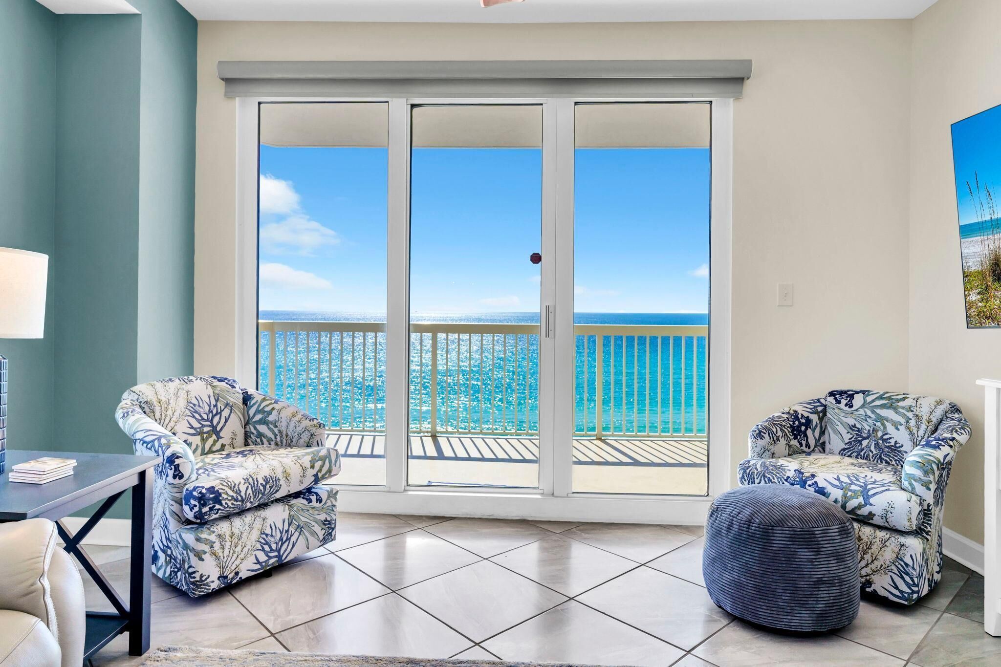 A room with a view of the ocean through a sliding glass door, two patterned armchairs, a small ottoman, and a table with books.
