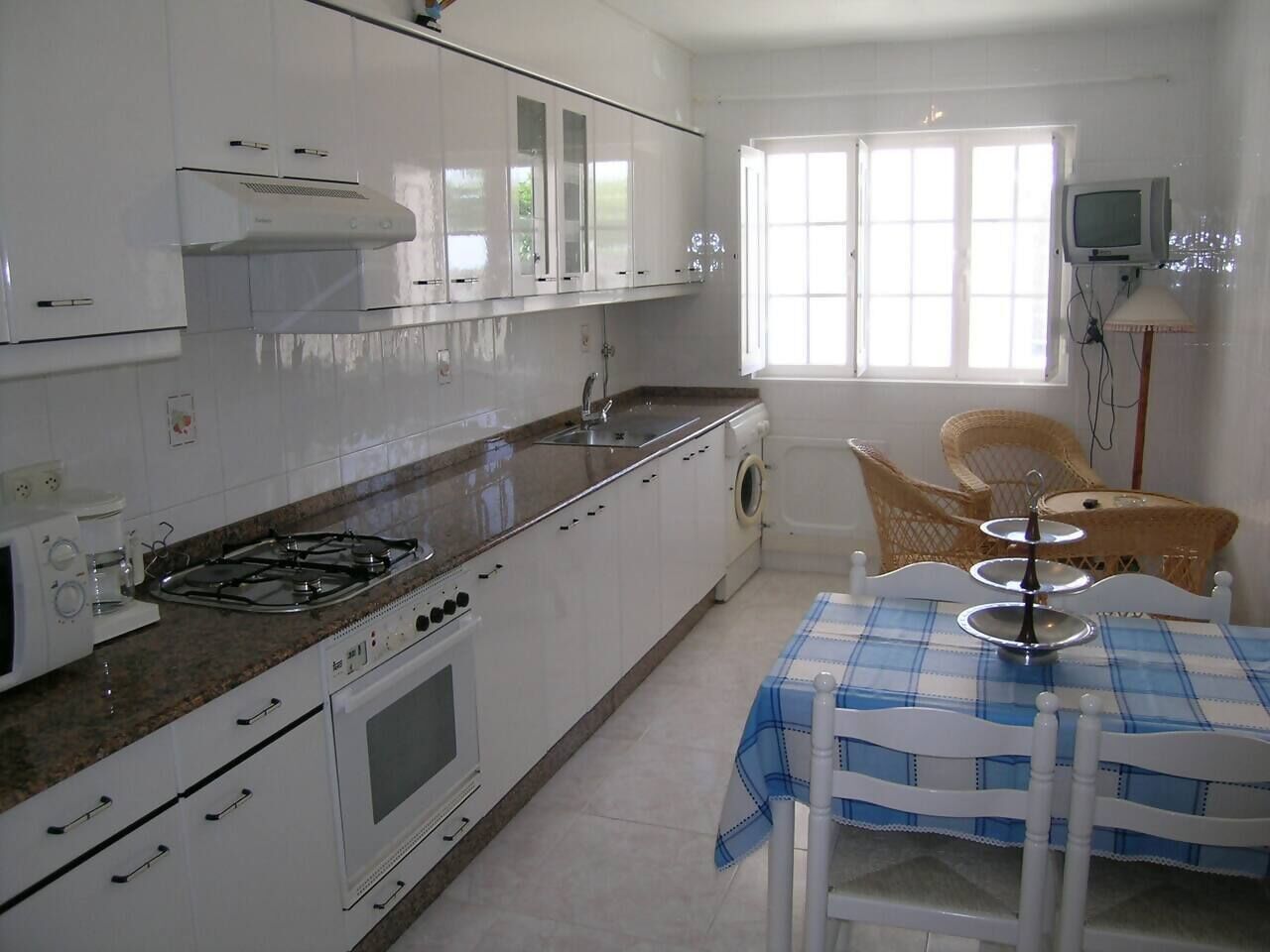 Une cuisine compacte avec des meubles blancs, une cuisinière à gaz, un four à micro-ondes et un lave-linge. On y trouve également un coin repas avec une table et des chaises, ainsi qu’une fenêtre qui laisse entrer la lumière naturelle.