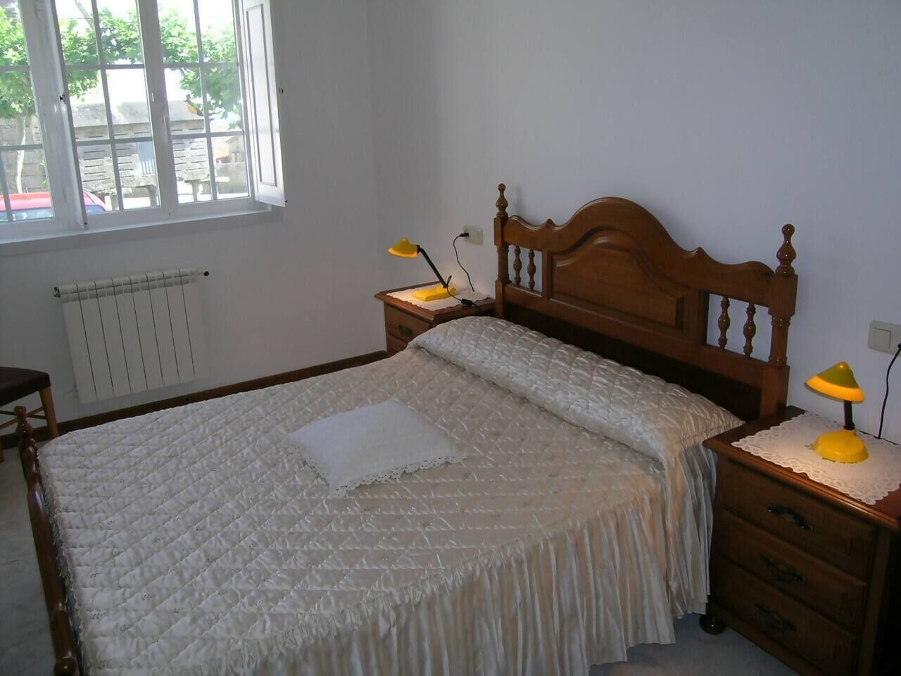 Un dormitorio con una cama de madera, dos mesitas de noche con lámparas, un radiador y una ventana con vistas a la calle.