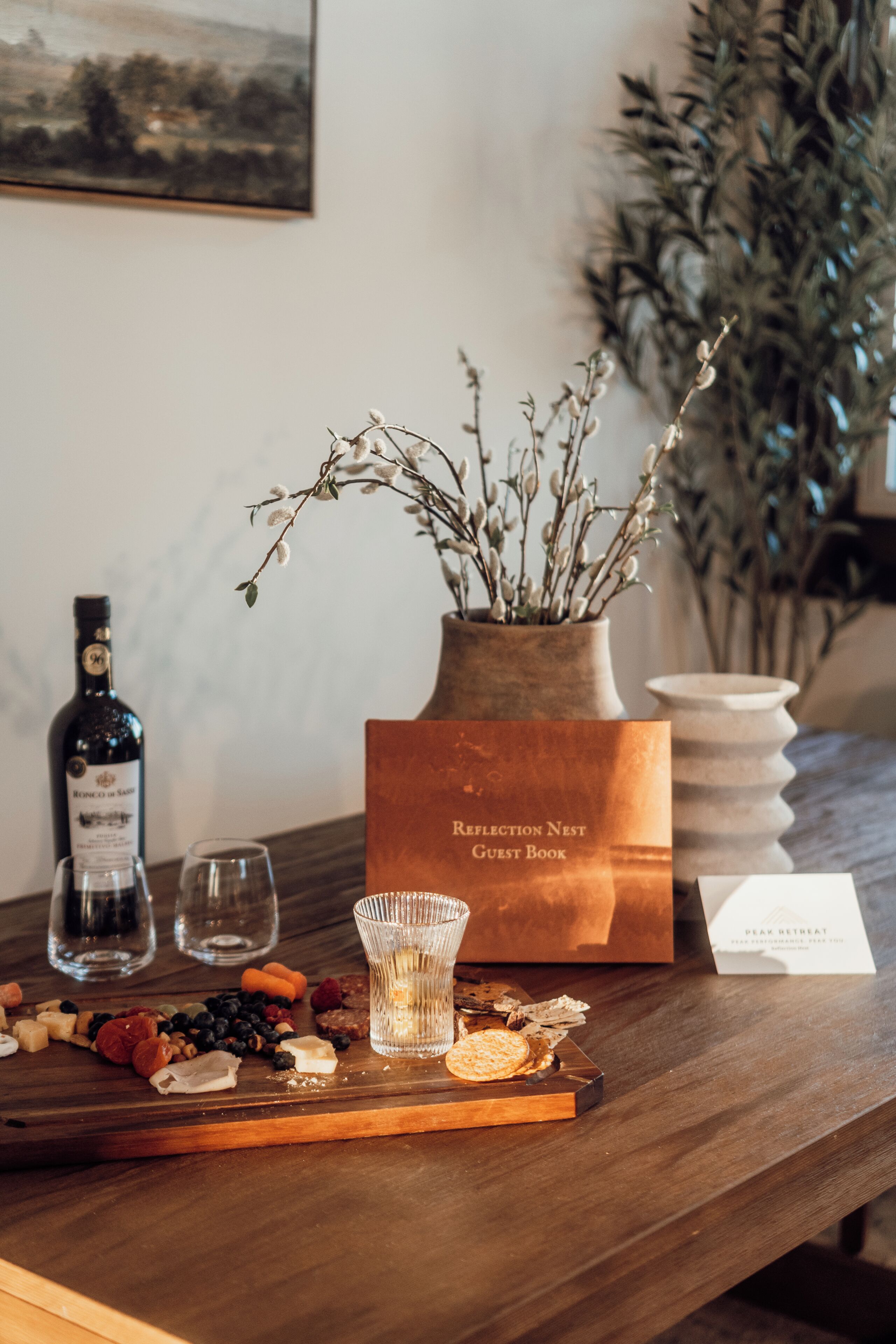 A wooden table with a bottle of wine, glasses, a tray with snacks, a guest book, and a vase with branches.