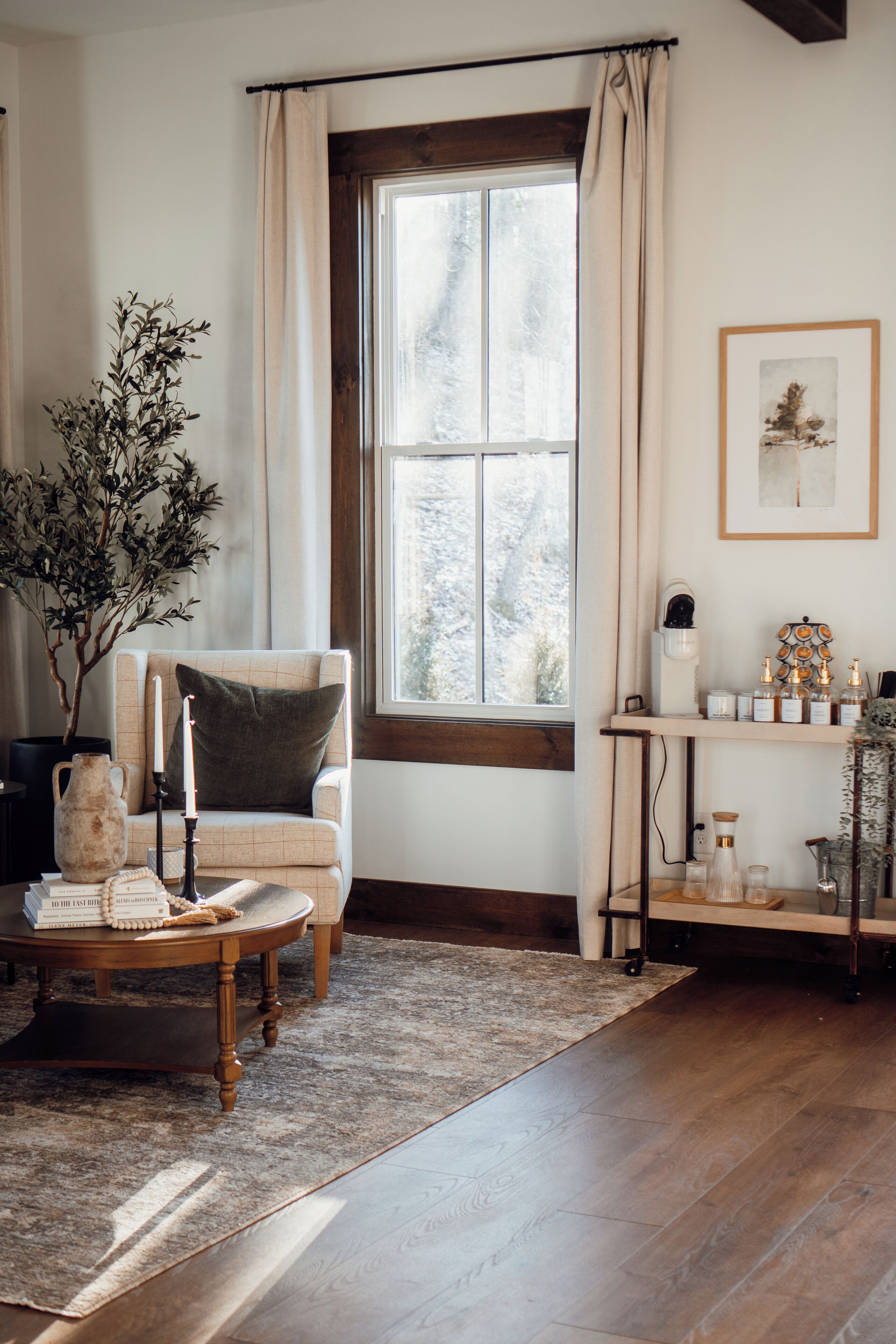 A cozy living room with a wooden floor, a comfortable armchair, a coffee table with books and a vase, a window with curtains, and a shelf with decorative items.