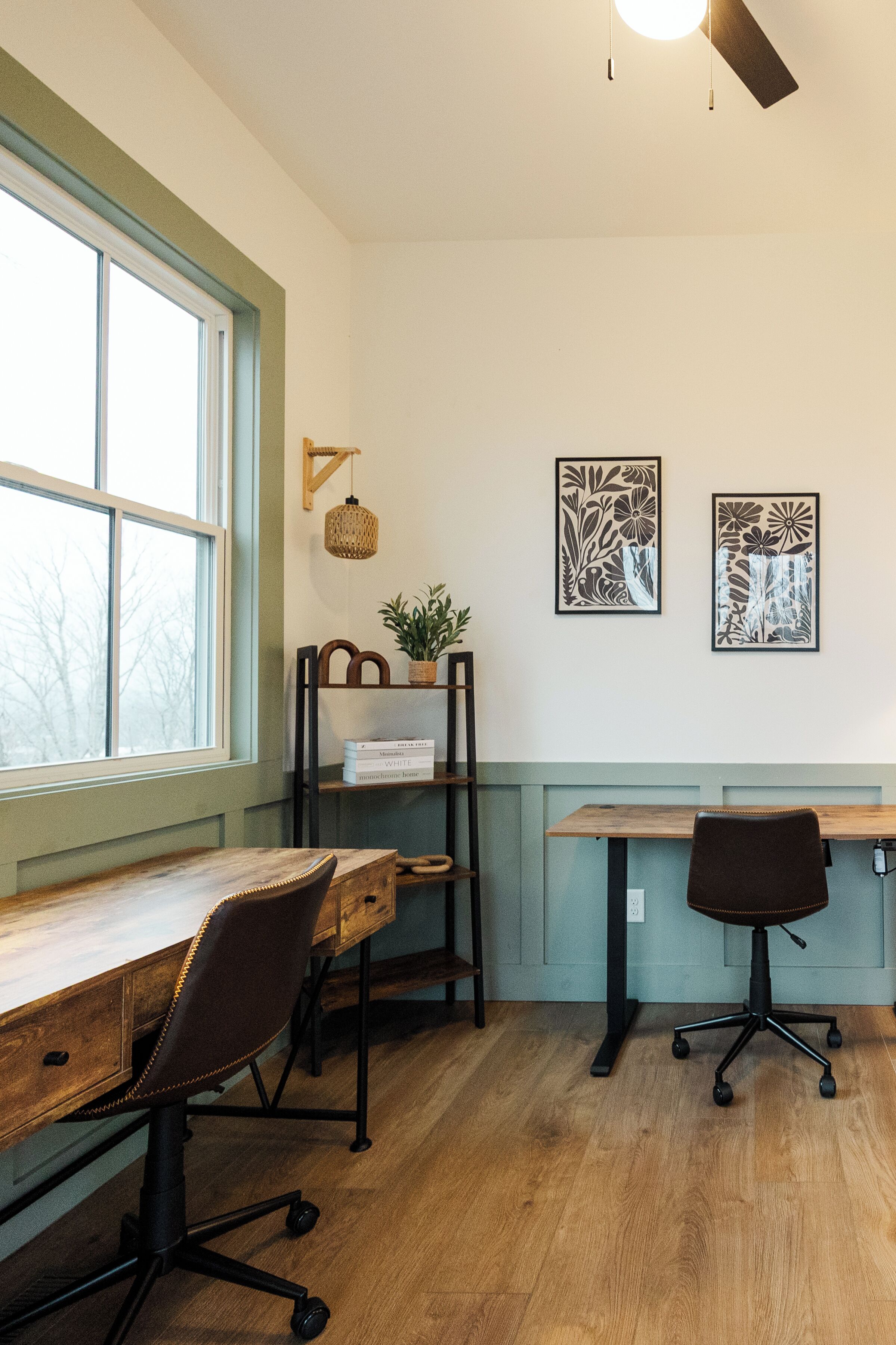 A room with a wooden desk, two chairs, a window with a view, a ceiling fan, and two wall-mounted artworks.