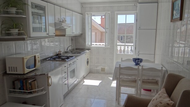 A compact kitchen with white cabinets, a microwave, a stove, and a sink. There's a small dining table with a blue bowl. - Ignacio C at the beach in Lariño (Lariño)