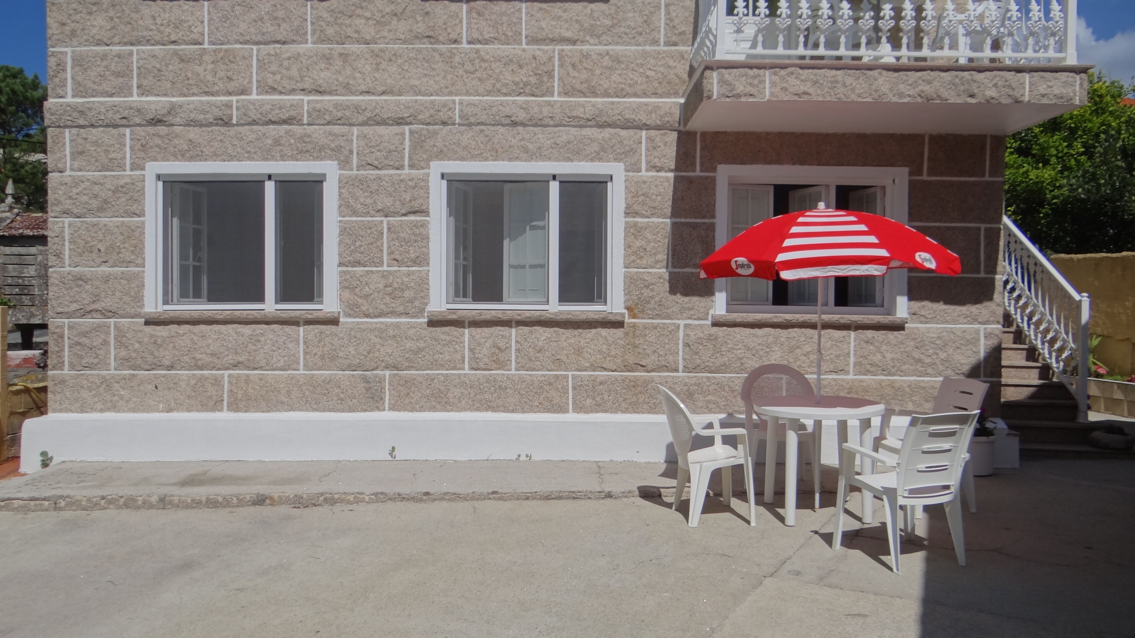 Un edificio de piedra con dos ventanas y un balcón. Se ve un paraguas blanco y rojo a rayas y sillas de plástico blancas en el exterior.