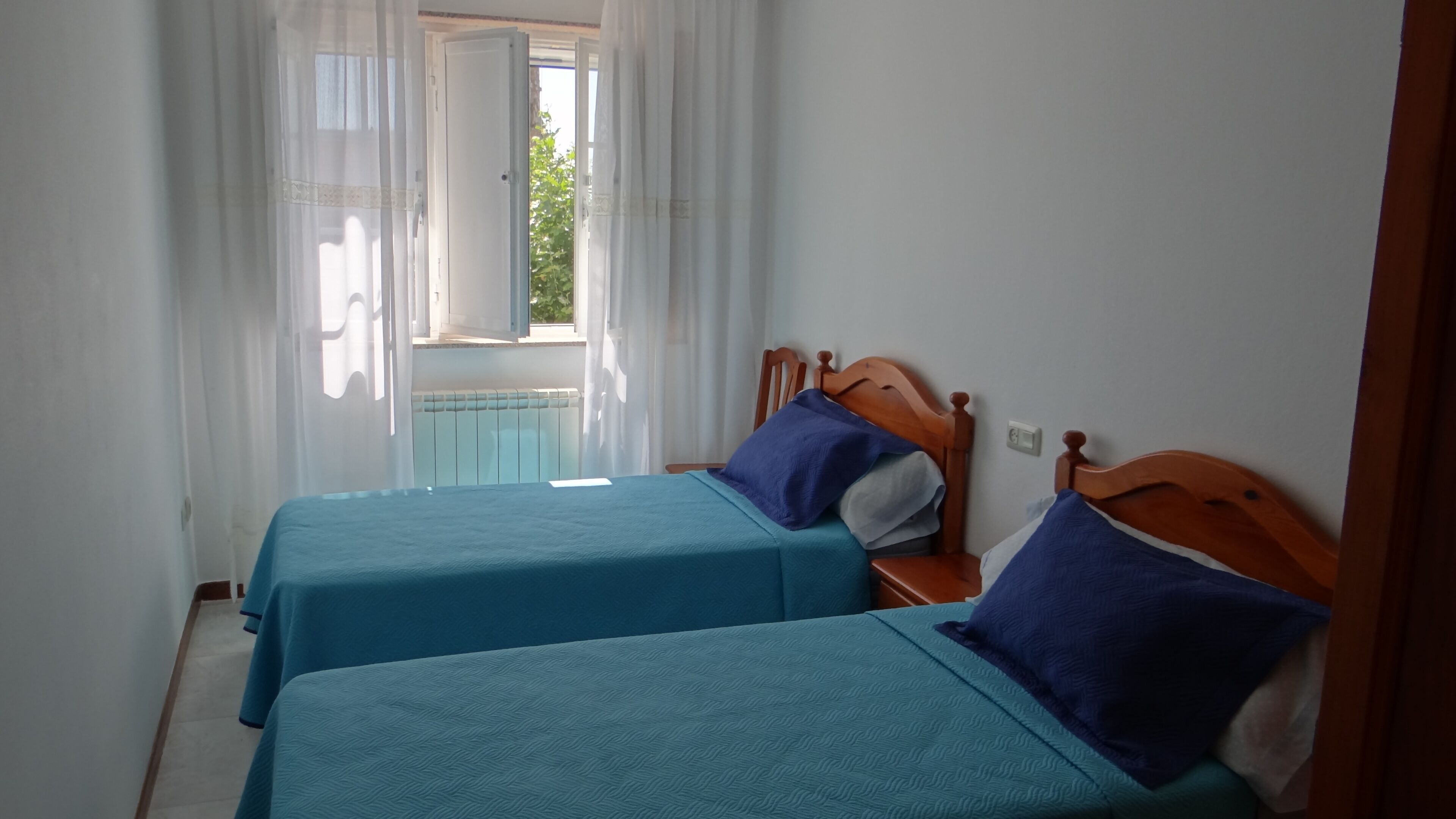 Una habitación con dos camas individuales, cabecero de madera, mesita de noche y ventana con cortinas de gasa.