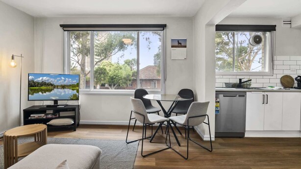 Una sala de estar moderna con un televisor de pantalla plana, un comedor con una mesa de vidrio y sillas, y una cocina con armarios blancos y un lavavajillas.